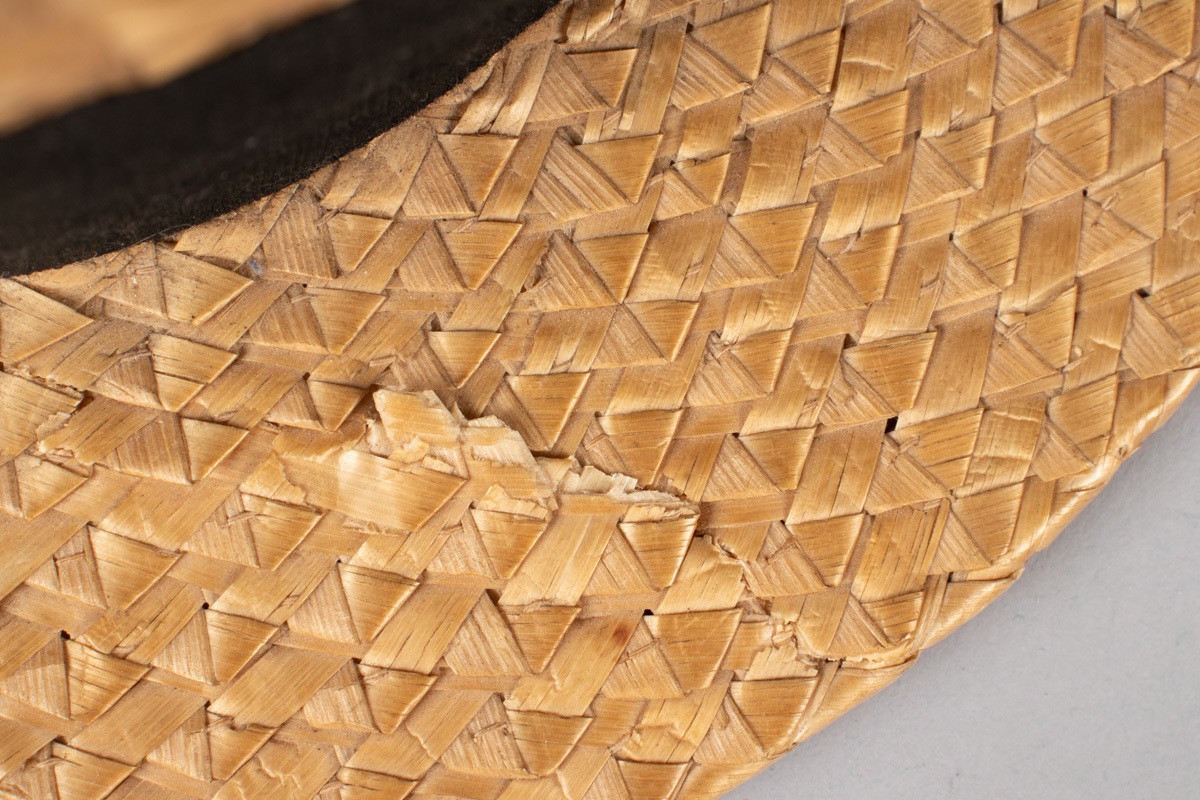 Stråhatten har en flat rett pull og bred flat brem. Svart bånd rundt pullen med fast sydd sløyfe. Svettebånd i lær og netting fôr. I bunnen er det festet et tøystykke med innlegg og logo. Bremen har fått en knekk og hattebåndet har en rift. Halmen er flettet med taggemønster. 