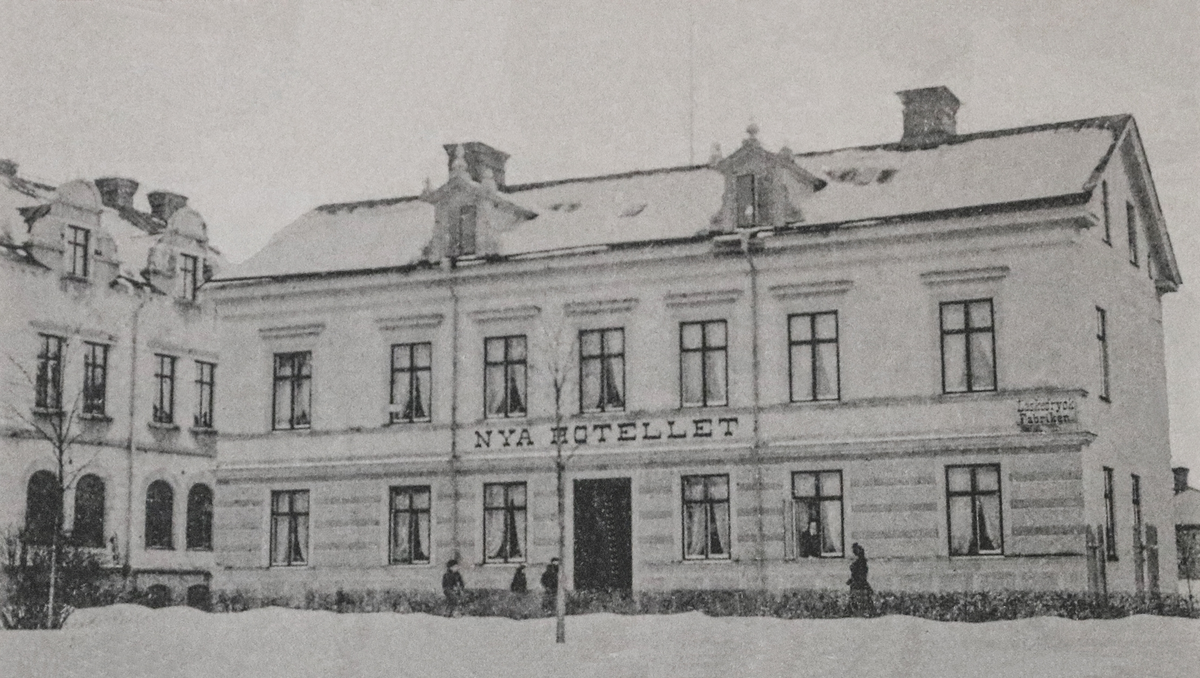 Vykort med motiv från Kyrkogatan, Enköping.  Vykortet visar "Nya Hotellet" på Kyrkogatan 5. Huset lät byggas omkring 1890 av plåtslagare/fabrikör B. Julius Åborg (1855-1934). I september 1892 öppnade han hotell för resande i nedervåningen under det nämnda hotellnamnet. På husets högra hörn syns en skylt med texten "Läskedrycksfabriken". Fotot är taget omkring 1900.