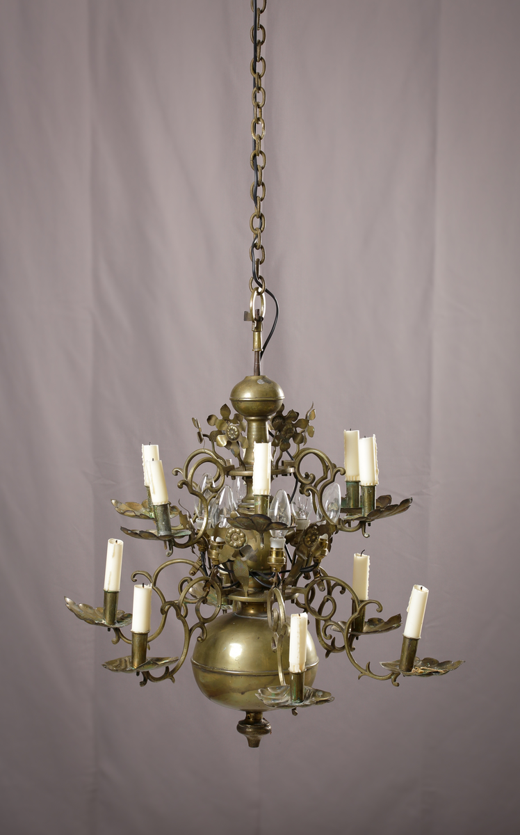 En ljuskrona av mässing, tolvarmad och försedd med anbringbara ornamenter. Kronan har profilerad mittstång, ändklot och tolv s-formade ljusarmar med cylindriska pipor och musselformade droppskålar. Reflexornamenten är blomformade. Ljuskronan är försedd med elektrisk belysning.

Inskrivet i huvudboken 1907.