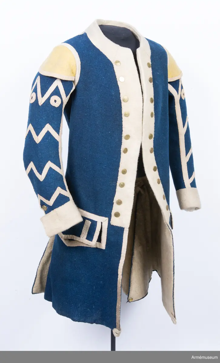 Rock av blått kläde med vita ärmuppslag och bröstrevärer av vitt kläde, gula axelkarmar, dekorationer av vävda vita band i tre olika bredder och släta mässingsknappar. Rocken är lång, med svängd mitt-fram-linje. Troligen nådde den nästan till knät på den som bar den. Den har 20 parvis ställda knappar fram, men knäpps kant i kant med två hyskor och hakar.
Den är konstruerad med hela framstycken som går runt till baksidan och bildar sidstycken. Delat bakstycke med mitt-bak-söm, samt tvåsömsärm. Sidsömmarna, ärmsömmarna och mitt-bak-sömmen är dekorerade med band. Två lågt ställda horisontella fickor fram med bandprydda ficklock, falska knapphål och två knappar vardera.
Skört med uppvik nertill fram. Sicksackdekorationer och rundlar på ärmarna.
Foder av vit boj i bålen, men oblekt bomull i ärmarna.