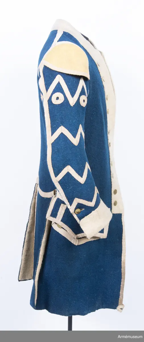 Rock av blått kläde med vita ärmuppslag och bröstrevärer av vitt kläde, gula axelkarmar, dekorationer av vävda vita band i tre olika bredder och släta mässingsknappar. Rocken är lång, med svängd mitt-fram-linje. Troligen nådde den nästan till knät på den som bar den. Den har 20 parvis ställda knappar fram, men knäpps kant i kant med två hyskor och hakar.
Den är konstruerad med hela framstycken som går runt till baksidan och bildar sidstycken. Delat bakstycke med mitt-bak-söm, samt tvåsömsärm. Sidsömmarna, ärmsömmarna och mitt-bak-sömmen är dekorerade med band. Två lågt ställda horisontella fickor fram med bandprydda ficklock, falska knapphål och två knappar vardera.
Skört med uppvik nertill fram. Sicksackdekorationer och rundlar på ärmarna.
Foder av vit boj i bålen, men oblekt bomull i ärmarna.