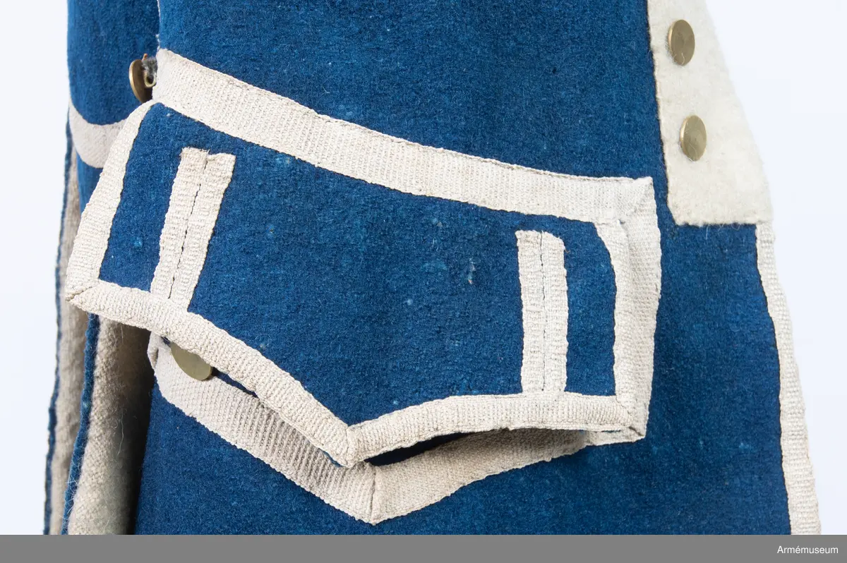 Rock av blått kläde med vita ärmuppslag och bröstrevärer av vitt kläde, gula axelkarmar, dekorationer av vävda vita band i tre olika bredder och släta mässingsknappar. Rocken är lång, med svängd mitt-fram-linje. Troligen nådde den nästan till knät på den som bar den. Den har 20 parvis ställda knappar fram, men knäpps kant i kant med två hyskor och hakar.
Den är konstruerad med hela framstycken som går runt till baksidan och bildar sidstycken. Delat bakstycke med mitt-bak-söm, samt tvåsömsärm. Sidsömmarna, ärmsömmarna och mitt-bak-sömmen är dekorerade med band. Två lågt ställda horisontella fickor fram med bandprydda ficklock, falska knapphål och två knappar vardera.
Skört med uppvik nertill fram. Sicksackdekorationer och rundlar på ärmarna.
Foder av vit boj i bålen, men oblekt bomull i ärmarna.