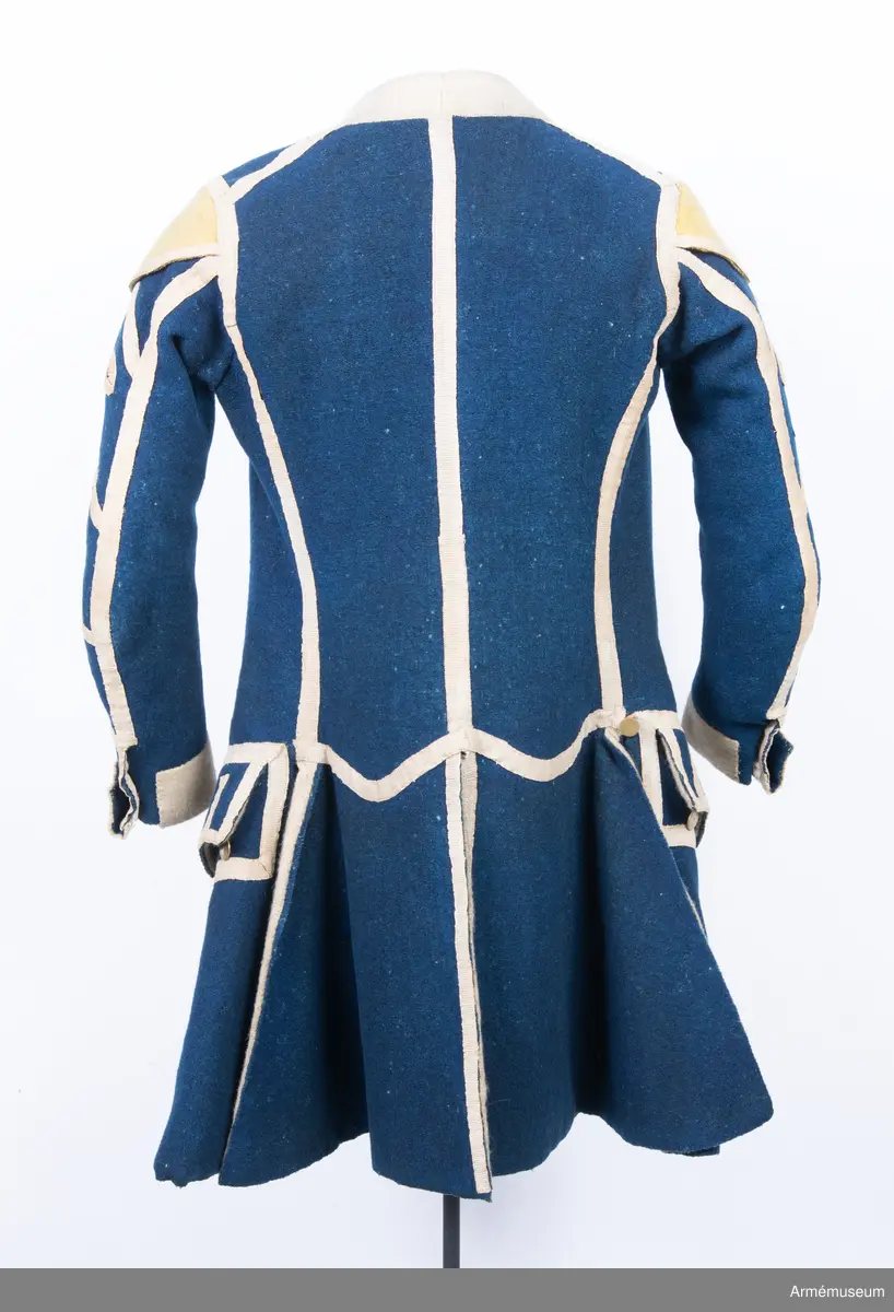 Rock av blått kläde med vita ärmuppslag och bröstrevärer av vitt kläde, gula axelkarmar, dekorationer av vävda vita band i tre olika bredder och släta mässingsknappar. Rocken är lång, med svängd mitt-fram-linje. Troligen nådde den nästan till knät på den som bar den. Den har 20 parvis ställda knappar fram, men knäpps kant i kant med två hyskor och hakar.
Den är konstruerad med hela framstycken som går runt till baksidan och bildar sidstycken. Delat bakstycke med mitt-bak-söm, samt tvåsömsärm. Sidsömmarna, ärmsömmarna och mitt-bak-sömmen är dekorerade med band. Två lågt ställda horisontella fickor fram med bandprydda ficklock, falska knapphål och två knappar vardera.
Skört med uppvik nertill fram. Sicksackdekorationer och rundlar på ärmarna.
Foder av vit boj i bålen, men oblekt bomull i ärmarna.