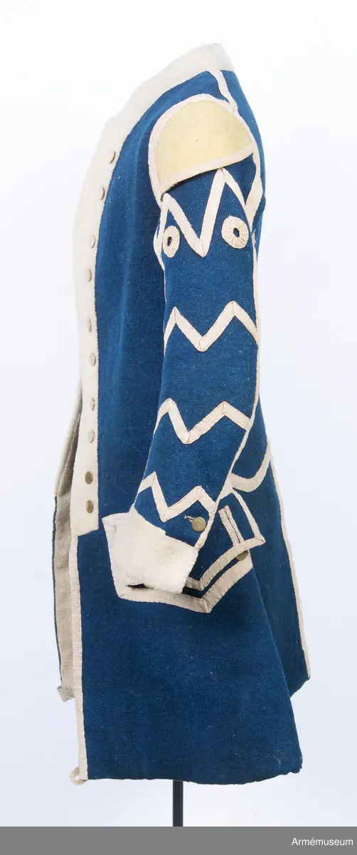 Rock av blått kläde med vita ärmuppslag och bröstrevärer av vitt kläde, gula axelkarmar, dekorationer av vävda vita band i tre olika bredder och släta mässingsknappar. Rocken är lång, med svängd mitt-fram-linje. Troligen nådde den nästan till knät på den som bar den. Den har 20 parvis ställda knappar fram, men knäpps kant i kant med två hyskor och hakar.
Den är konstruerad med hela framstycken som går runt till baksidan och bildar sidstycken. Delat bakstycke med mitt-bak-söm, samt tvåsömsärm. Sidsömmarna, ärmsömmarna och mitt-bak-sömmen är dekorerade med band. Två lågt ställda horisontella fickor fram med bandprydda ficklock, falska knapphål och två knappar vardera.
Skört med uppvik nertill fram. Sicksackdekorationer och rundlar på ärmarna.
Foder av vit boj i bålen, men oblekt bomull i ärmarna.