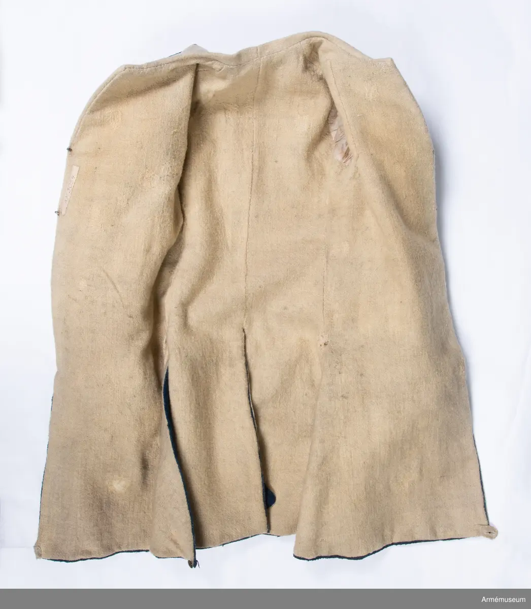 Rock av blått kläde med vita ärmuppslag och bröstrevärer av vitt kläde, gula axelkarmar, dekorationer av vävda vita band i tre olika bredder och släta mässingsknappar. Rocken är lång, med svängd mitt-fram-linje. Troligen nådde den nästan till knät på den som bar den. Den har 20 parvis ställda knappar fram, men knäpps kant i kant med två hyskor och hakar.
Den är konstruerad med hela framstycken som går runt till baksidan och bildar sidstycken. Delat bakstycke med mitt-bak-söm, samt tvåsömsärm. Sidsömmarna, ärmsömmarna och mitt-bak-sömmen är dekorerade med band. Två lågt ställda horisontella fickor fram med bandprydda ficklock, falska knapphål och två knappar vardera.
Skört med uppvik nertill fram. Sicksackdekorationer och rundlar på ärmarna.
Foder av vit boj i bålen, men oblekt bomull i ärmarna.