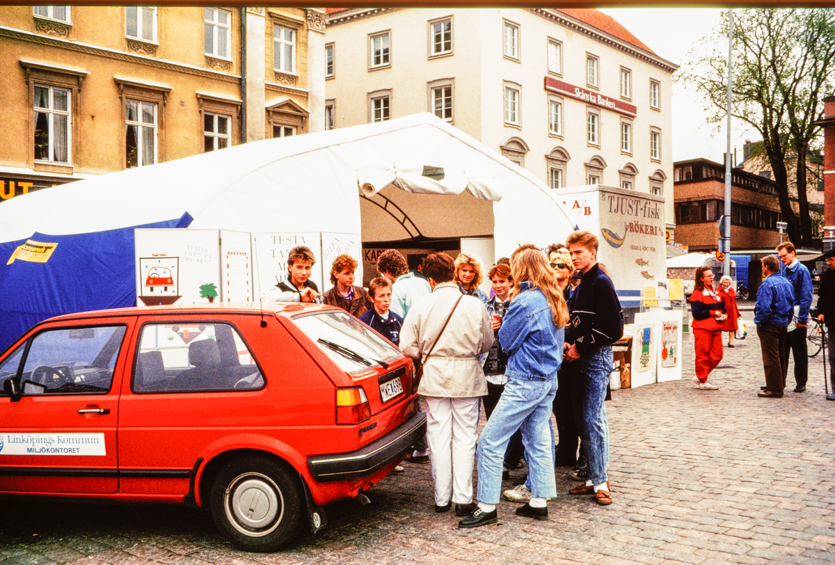 Stora torget i Linköping. Utställning och handel på torget.
Man ser ungdomar och en bil märkt Miljökontoret i Linköpings kommun. Bilder från staden Linköping digitaliserade från diapositiv. Bilderna är från 1970-1990-talet.