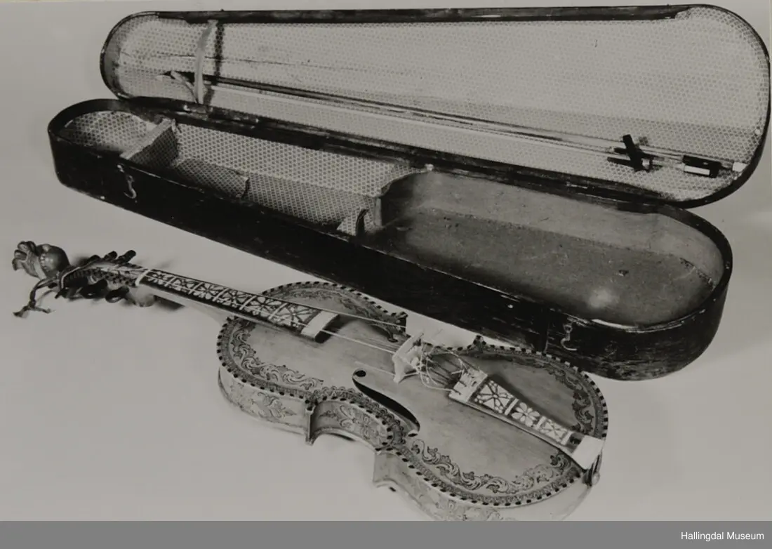 Hardingfele. Fela er litt kuven på midten, og har to S-forma lydhol i loket. Gripebrettet er dekorert med innfelte bitar av perlemor, og det er skore eit dyrehovud med krone i enden av halsen. Fela er dekorert langs kanten, med blomedekor. 