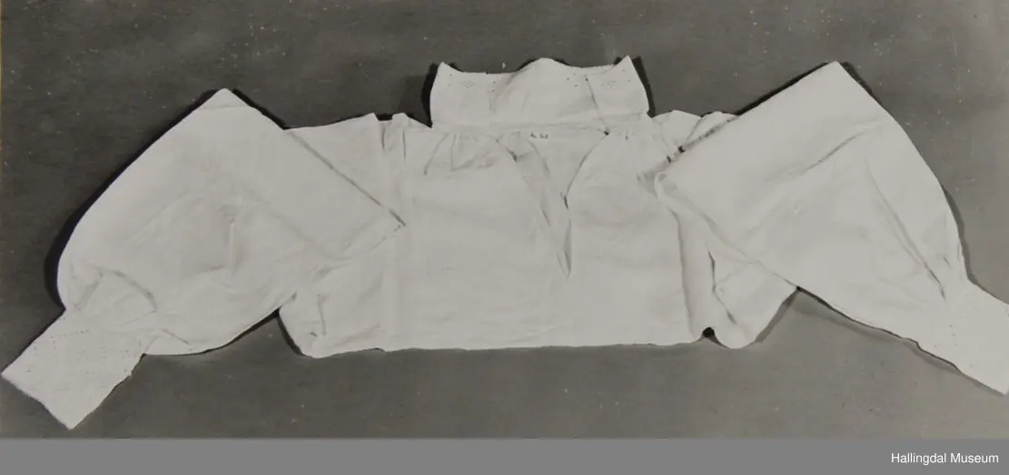 Rynket til halslinning. Kile ved skulder, rynket til halslinning, Erme rett ved skulder, rynket til mansjett. Kile under erme. Broderi i kvitsaum på linninger.