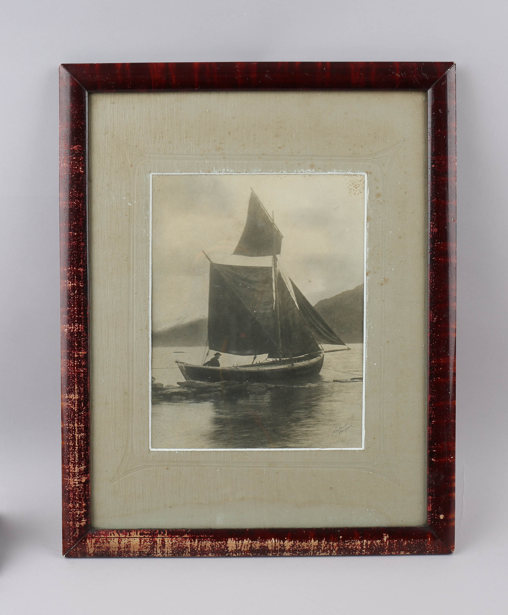 Svart-kvitt fotografi i glas og ramme. Motivet er ein båt med full seglføring, ein person ved rorstanga. Rundt motivet er det passepartout med ein prega bord. Ramma er mahognyfarga, med svakt konveks profil.