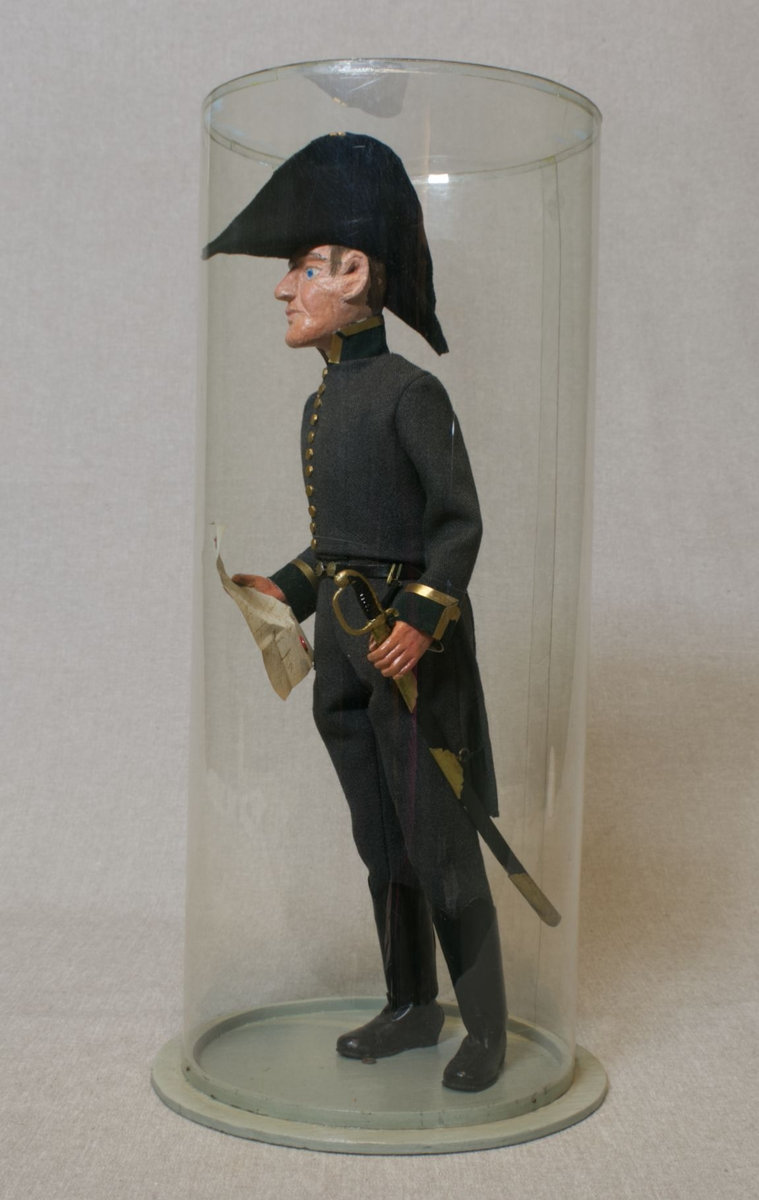Grå kjole og benklær, grønn krage og oppslag, sorte støvler, sort snutehatt med gullkrampe, sabel med geheng. Montert på sirkelrund plate. Modellen har gradstegn for politiadjutant. Modellen står inni en plastsylinder på en treplate.