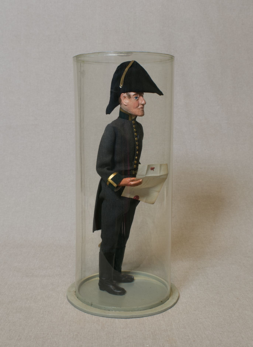 Grå kjole og benklær, grønn krage og oppslag, sorte støvler, sort snutehatt med gullkrampe, sabel med geheng. Montert på sirkelrund plate. Modellen har gradstegn for politiadjutant. Modellen står inni en plastsylinder på en treplate.