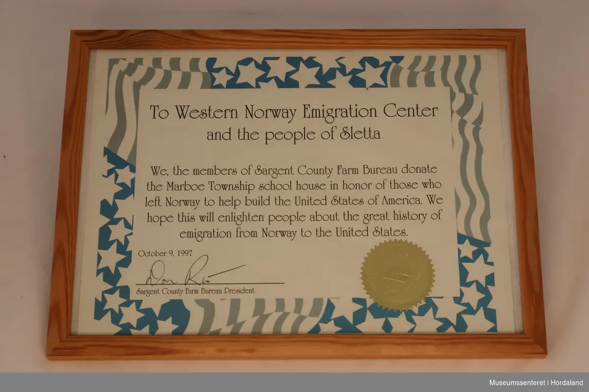 Innrammet donasjonsbevis. Det har liggende, rektangulær form, er av papir bak glass med treramme. Papiret beskriver at Sargent County Farm Bureau donerte Skulestova til Vestnorsk utvandringsenter i 1997. Det er dekorert med det amerikanske flagget i blått, hvitt og grått, og har et rundt, gyllent stempel.