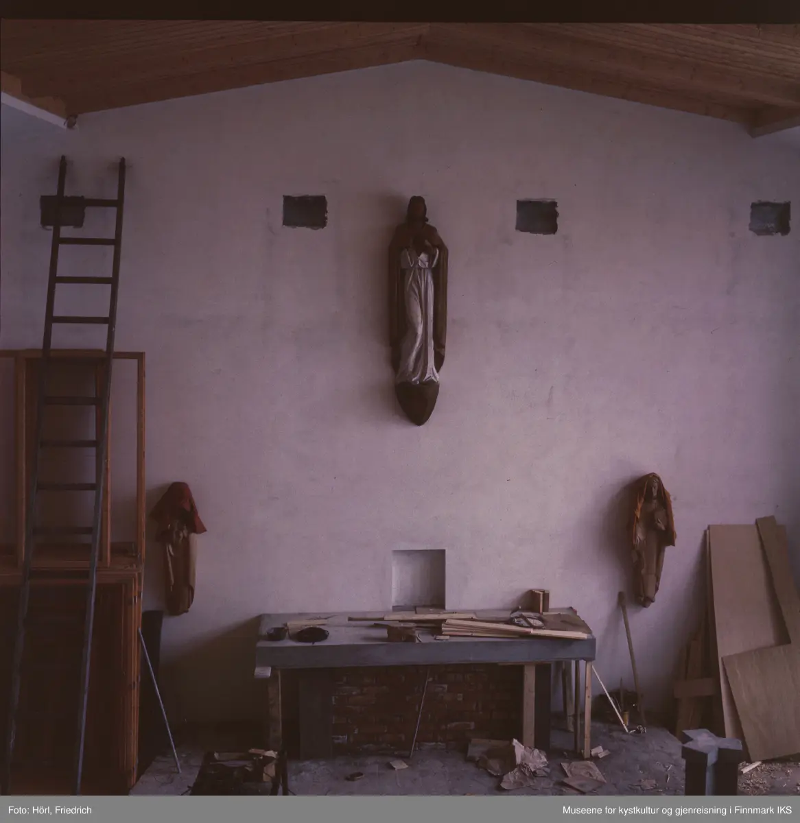 Fotografiet er noe underbelyst. Den viser interiøret av Den katolske kirke St. Mikael i Hammerfest i byggeperioden. Alteret er under byggingen. Skulpturene på veggen til venstre og høyre for det er dekket med tepper. Til venstre i bildet skimter man en lang stige som er lent mot veggen.