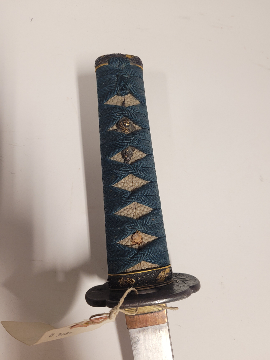 Samurai-dolk (WAKICACHI) från Japan. Klinga svagt bakåtböjd,eneggad, skarp, facetterad rygg. Fäste av vitt hajskinn flätat med grönt bomullsband med två amuletter, delvis förgyllda. Beslag upptill och nedtill cicilerade och förgyllda. Parerplåt rund med ett hål på ena sidan samt med förgyllt och cicilerat bladverk. Balja av trä, svartlackerad med beslag å fyra ställen.
Låsfjädern,d.v.s. bestickknivens handtag saknas.
Kniven på baljan (KOZUKA) saknas.
Från fregatten VANADIS världsomsegling 1883-85.