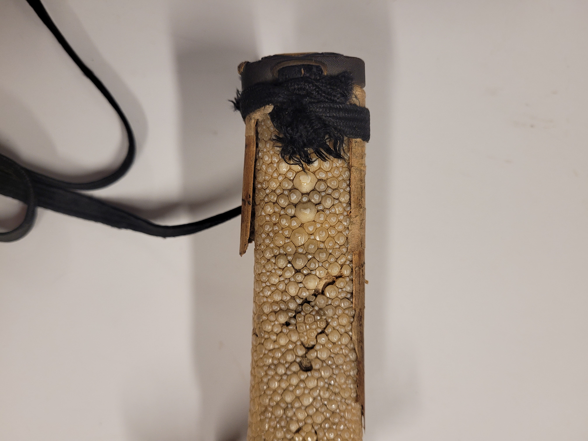Samurajsvärd från Japan. Klinga svagt bakåtböjd, eneggad skarp, facetterad rygg. Fäste av vitt hajskinn (stingrocka?), flätad med svart bomullsgarn samt försett med två amuletter, den ena med guldinläggningar. Fästets beslag upptill och nedtill har förgyllda inläggningar. Klinga märkt: Lindman. Parerplåt rund, genombrutet bladverk med fåglar.