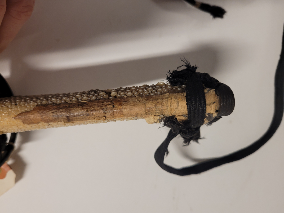 Samurajsvärd från Japan. Klinga svagt bakåtböjd, eneggad skarp, facetterad rygg. Fäste av vitt hajskinn (stingrocka?), flätad med svart bomullsgarn samt försett med två amuletter, den ena med guldinläggningar. Fästets beslag upptill och nedtill har förgyllda inläggningar. Klinga märkt: Lindman. Parerplåt rund, genombrutet bladverk med fåglar.