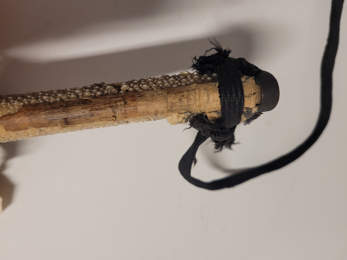 Samurajsvärd från Japan. Klinga svagt bakåtböjd, eneggad skarp, facetterad rygg. Fäste av vitt hajskinn (stingrocka?), flätad med svart bomullsgarn samt försett med två amuletter, den ena med guldinläggningar. Fästets beslag upptill och nedtill har förgyllda inläggningar. Klinga märkt: Lindman. Parerplåt rund, genombrutet bladverk med fåglar.