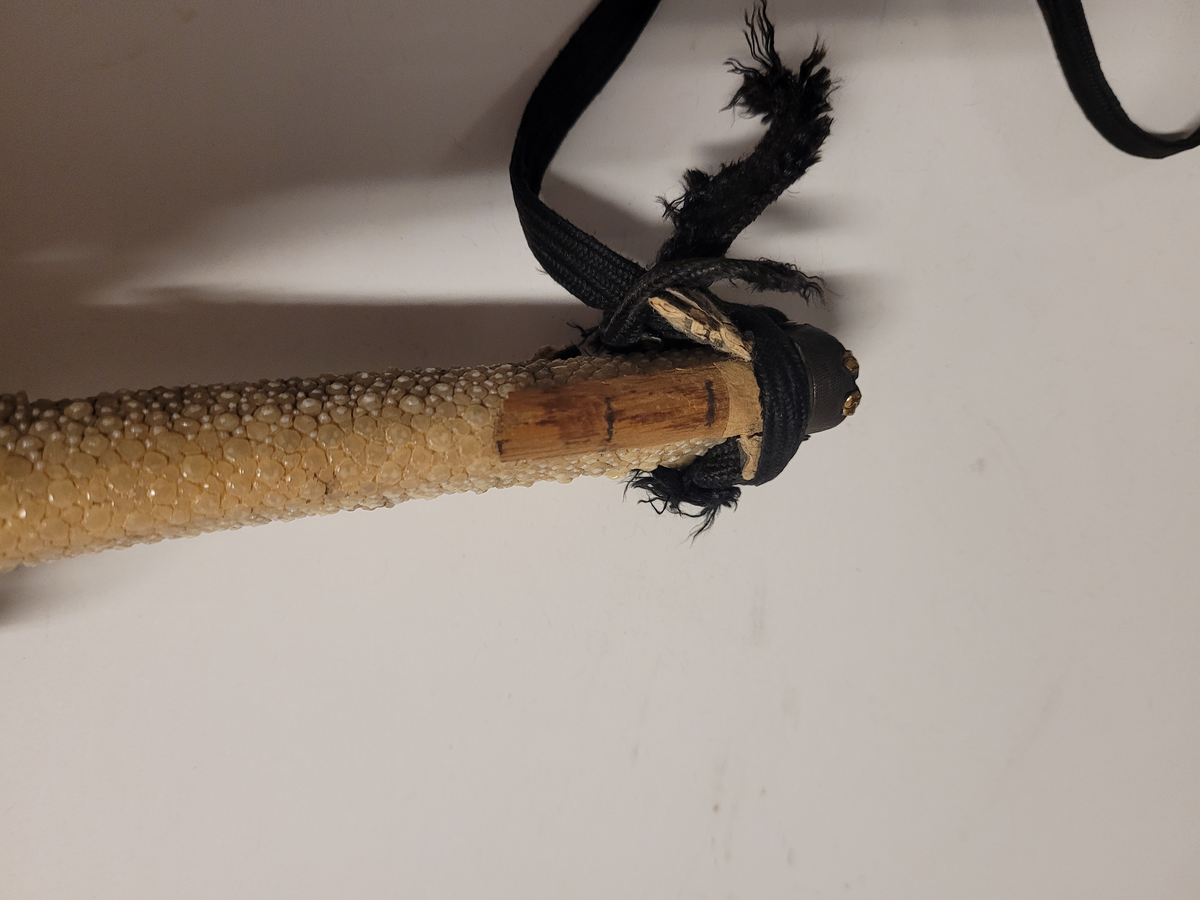 Samurajsvärd från Japan. Klinga svagt bakåtböjd, eneggad skarp, facetterad rygg. Fäste av vitt hajskinn (stingrocka?), flätad med svart bomullsgarn samt försett med två amuletter, den ena med guldinläggningar. Fästets beslag upptill och nedtill har förgyllda inläggningar. Klinga märkt: Lindman. Parerplåt rund, genombrutet bladverk med fåglar.