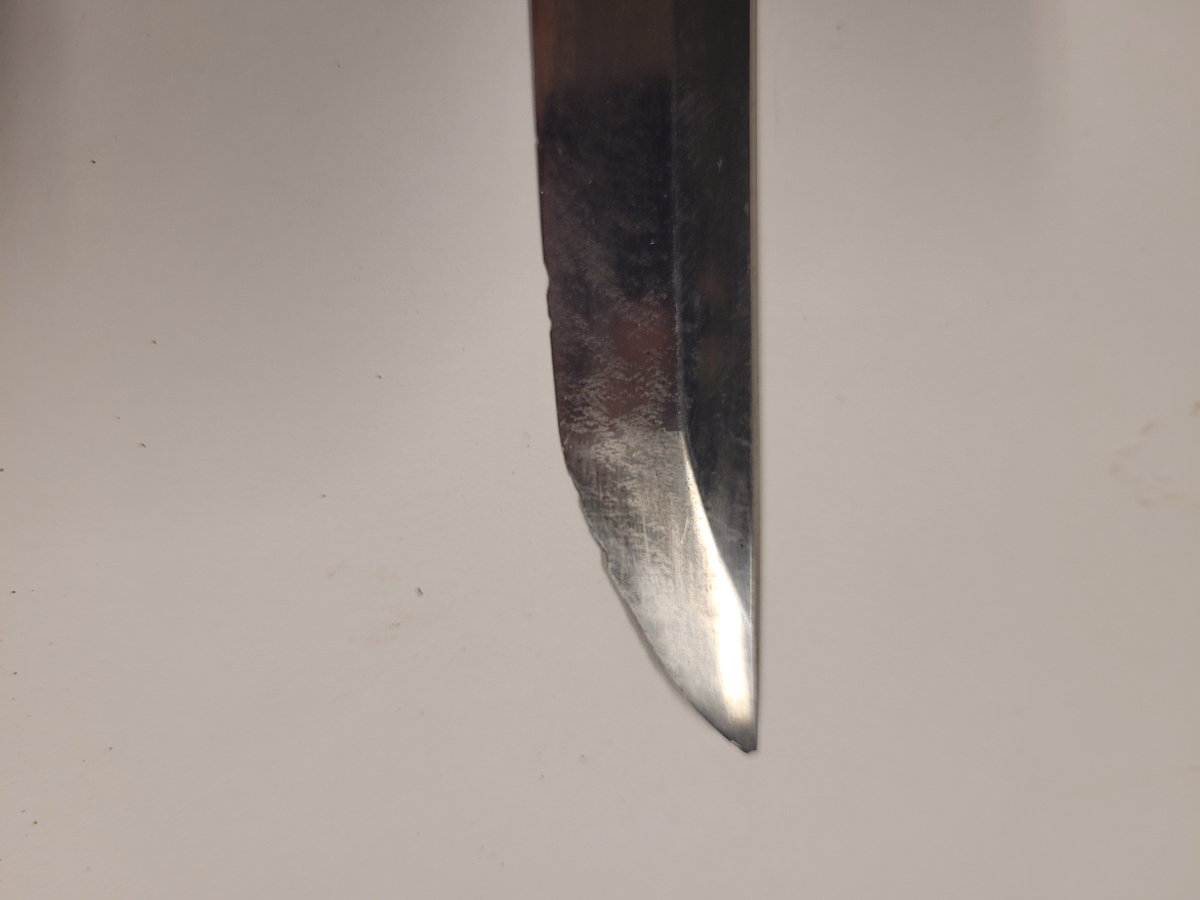 Samurajsvärd från Japan. Klinga svagt bakåtböjd, eneggad skarp, facetterad rygg. Fäste av vitt hajskinn (stingrocka?), flätad med svart bomullsgarn samt försett med två amuletter, den ena med guldinläggningar. Fästets beslag upptill och nedtill har förgyllda inläggningar. Klinga märkt: Lindman. Parerplåt rund, genombrutet bladverk med fåglar.