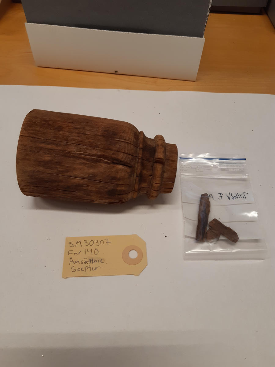 Ansättare som är snyggt svarvad. Hål för skaft mäter 27 mm. Trät som smör, delar av ytan faller sönder. Skadad på en sida. Rest av materialet i skafthålet undersöktes under arbetsmikroskop för att utesluta att det var något som tillhörde föremålet. Består av sediment med organiskt material. Prov sparat i påse.