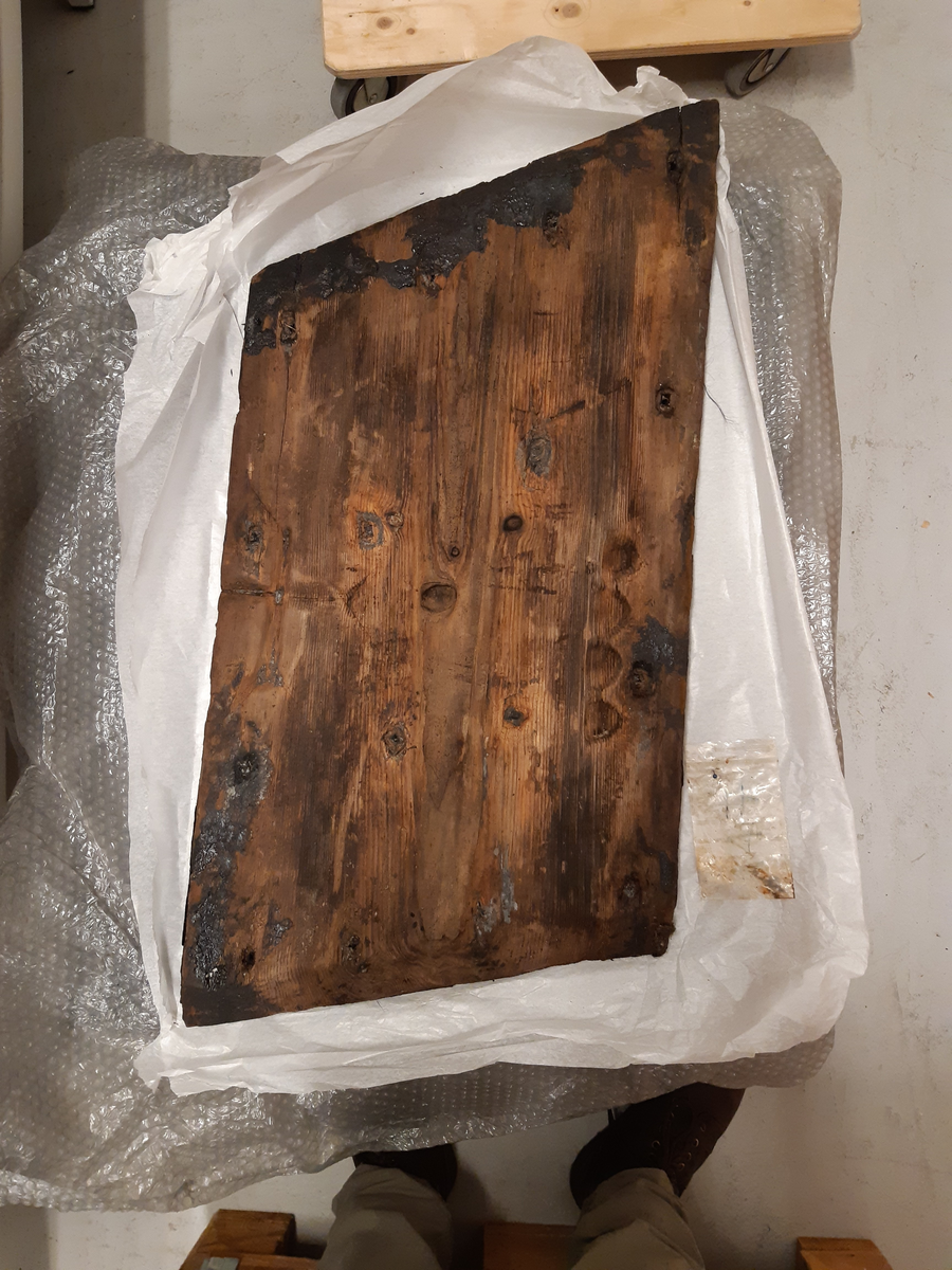 Del av fyrhydning eller extra skrov med rester av fibermatta. Möjligen rester av spik i spikhålen. Mycket järn i ytan på i synnerhet på ytan inåt med rester av fibrer.