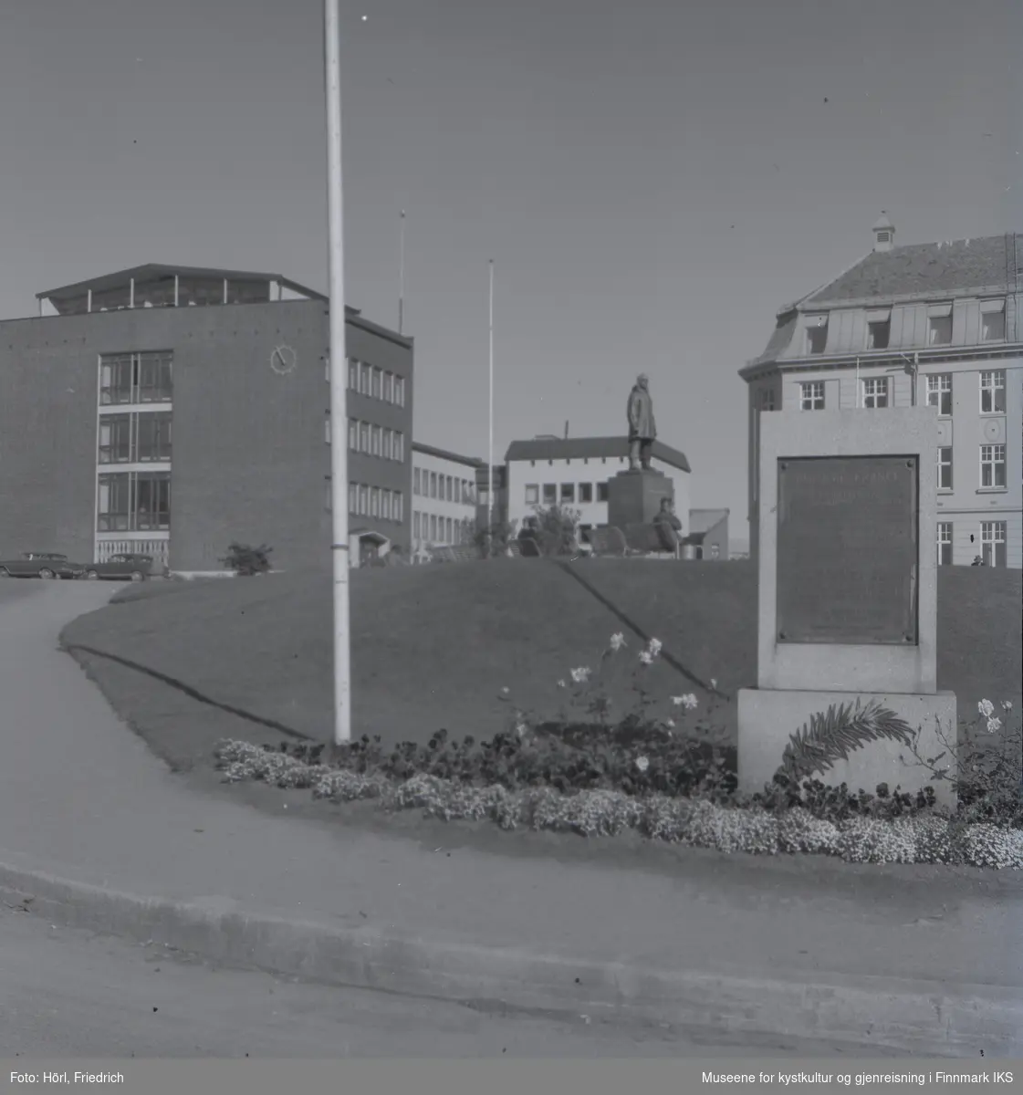 Lathammonumentet ved Roald Amundsen plass i Tromsø. I forgrunnen ser man monumentet som består av en stein med bronseplate. På sokkelen er det et metallornament som er utformet som en palmekvist. Rundt minnesmerket er det anlagt et blomsterbed. Lenger i bakgrunnen ser man bronsestatuen av Roald Amundsen. Personer sitter på benker rundt statuen.