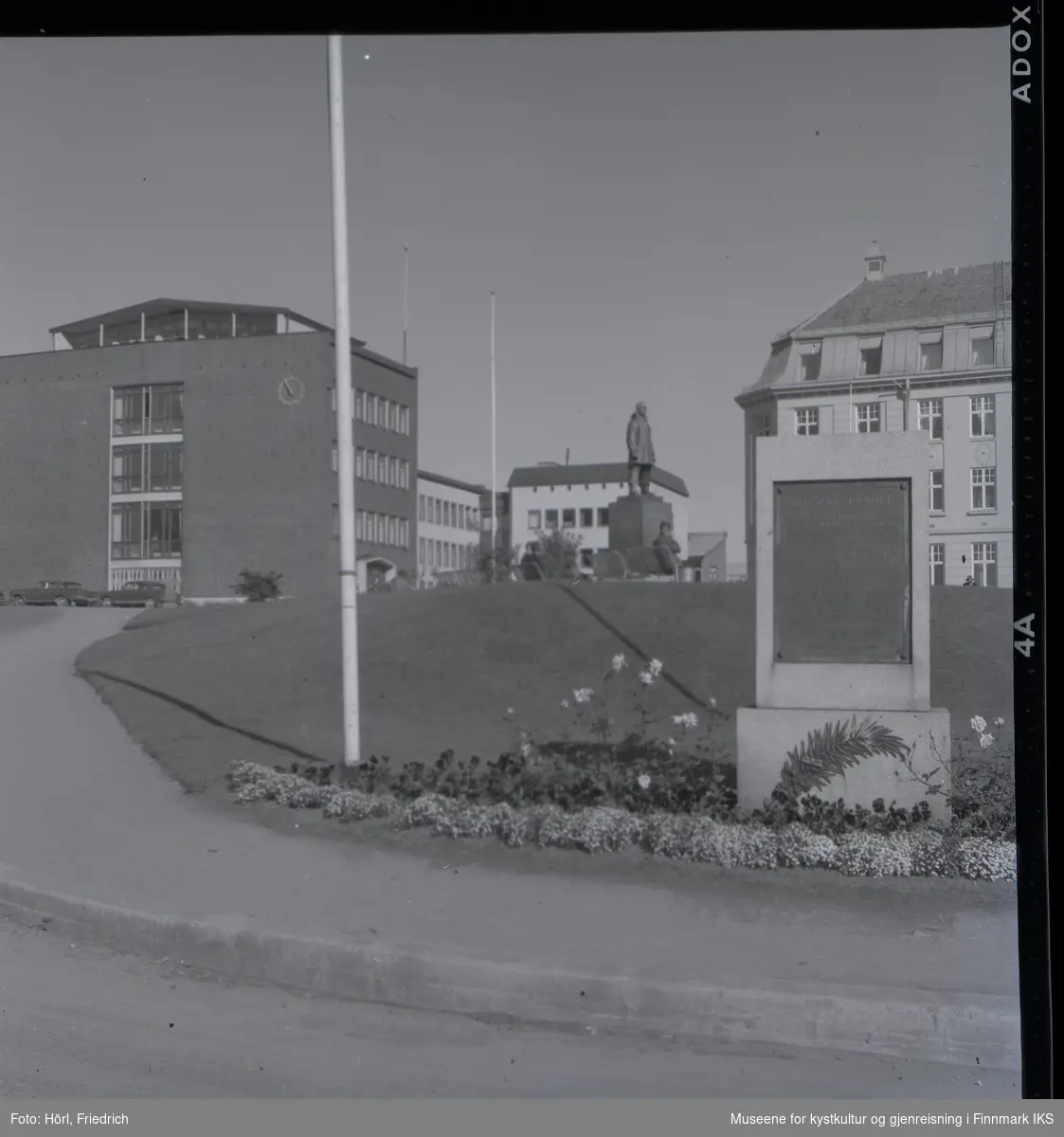 Lathammonumentet ved Roald Amundsen plass i Tromsø. I forgrunnen ser man monumentet som består av en stein med bronseplate. På sokkelen er det et metallornament som er utformet som en palmekvist. Rundt minnesmerket er det anlagt et blomsterbed. Lenger i bakgrunnen ser man bronsestatuen av Roald Amundsen. Personer sitter på benker rundt statuen.