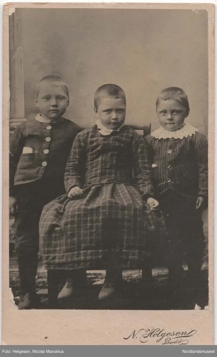 Portrett av tre barn, to gutter i rutet og stripet jakke, mellom dem ei jente i rutet kjole på en stol. Helfigur.