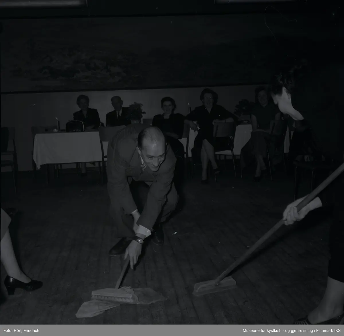 Bildet viser Theodor Trondsen og Petra Trondsen på juletrefest i den katolske menigheten i 1957. De ler mens de er med i en lek der de holder hver sin skrubb og tydeligvis kjemper om ei gulvklut. I bakgrunnen sitter flere personer som ser på og smiler. Bak dem på veggen er det et stort landskapsmaleri.