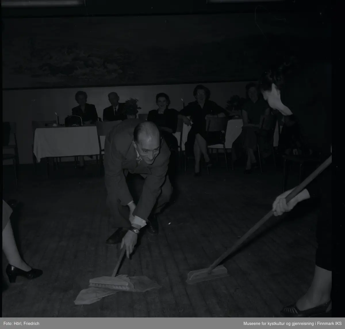 Bildet viser Theodor Trondsen og Petra Trondsen på juletrefest i den katolske menigheten i 1957. De ler mens de er med i en lek der de holder hver sin skrubb og tydeligvis kjemper om ei gulvklut. I bakgrunnen sitter flere personer som ser på og smiler. Bak dem på veggen er det et stort landskapsmaleri.