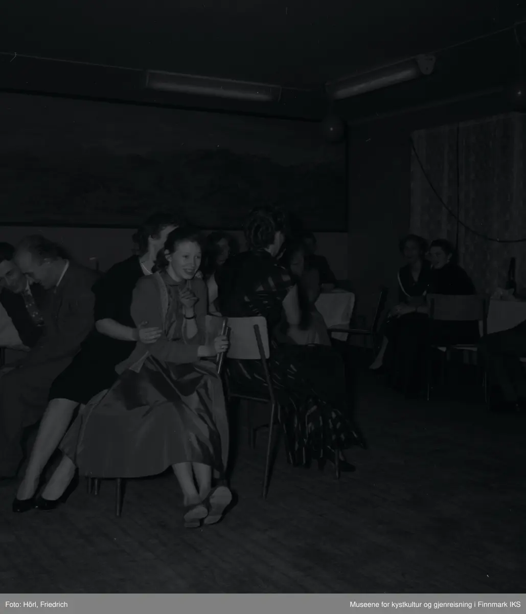 Den katolske menigheten i Hammerfest feirer juletrefest i 1957. De voksne på bildet spiller stolleken, også kjent som "Reisen til Jerusalem". Deltagerne og publikum er i god stemning. Rommet er pyntet med ballonger og på veggen i bakgrunnen er det et stort landskapsmaleri.