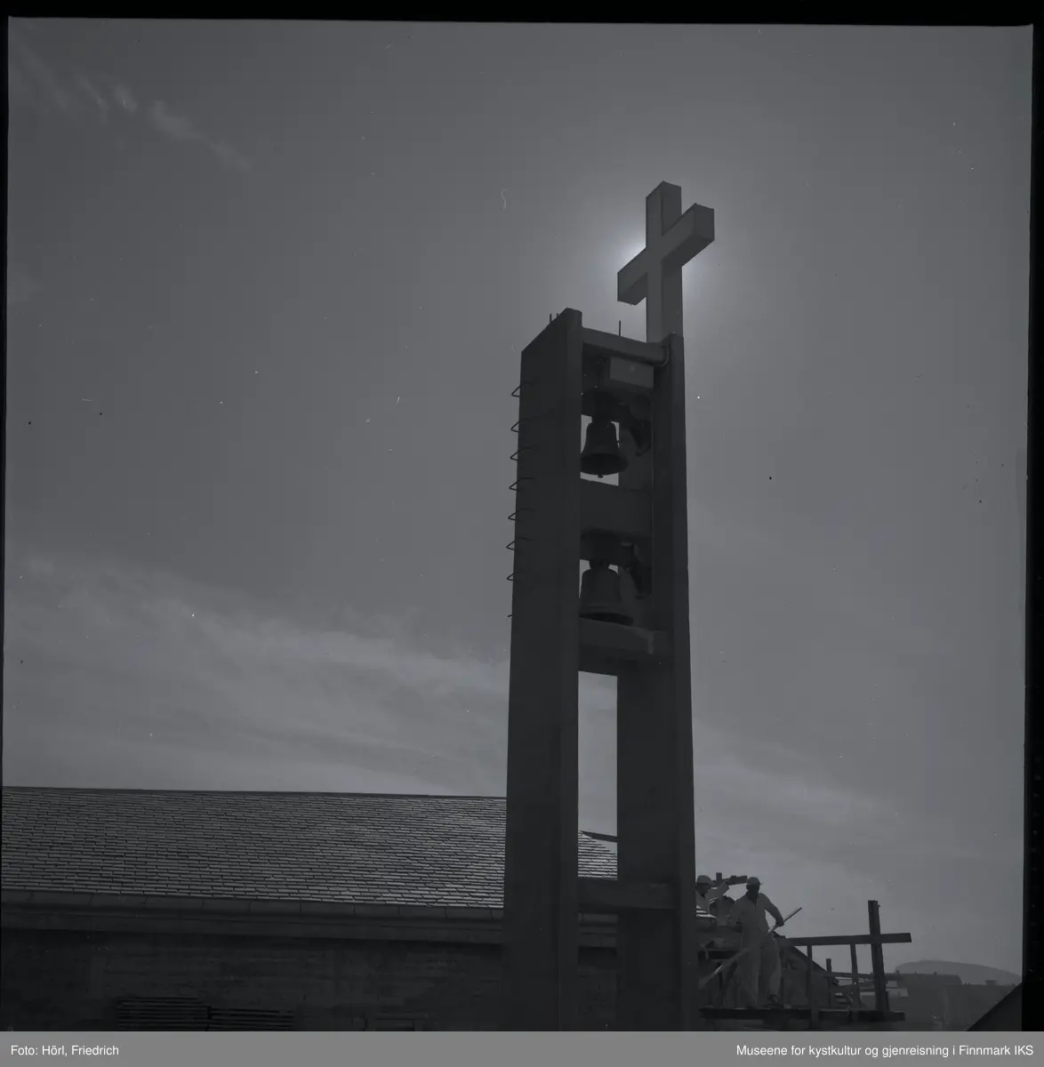 Bildet viser klokketårnet til den katolske kirka St. Mikael i Hammerfest i motlys av sola i 1958. Bildet er tatt fra sidebygget ut mot Sorøysundet der man ser Håja fjellet. Taket til kirka er ferdig bygd og belagt med skifer. På nedsida av kirka står det fortsatt et stillas og man kan se to arbeider der. I tårnet henger igjen de klokkene som allerede tilhørte til kirka før ødeleggelsene under andre verdenskrig. Det lyssatte korset er montert, men det er foreløpig kun sola som skinner.