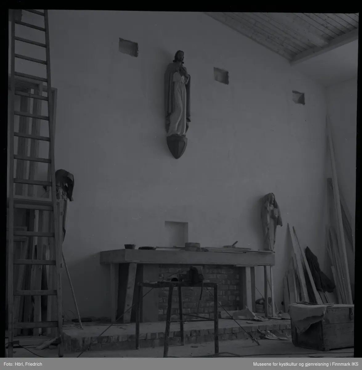 Interiøret av den katolske kirka St. Mikael i Hammerfest bygges i 1958. I motsetning til senere, er det montert en skulptur av Jesus Kristus ovenfor alteret. Senere ble det plassert et krusifiks der. Mosaikken på fronten av alteret, som ble utført av Karl Manninger, er ikke kommet på plass enda. Til venstre for alteret er det montert enn skulptur av Maria med barnet, til høyre vises Josef. Skulpturene, som er skjært av tre, er avdekket med tepper. Til venstre i bilde står en lang stige og til høyre noe mer byggemateriale lent mot veggen.