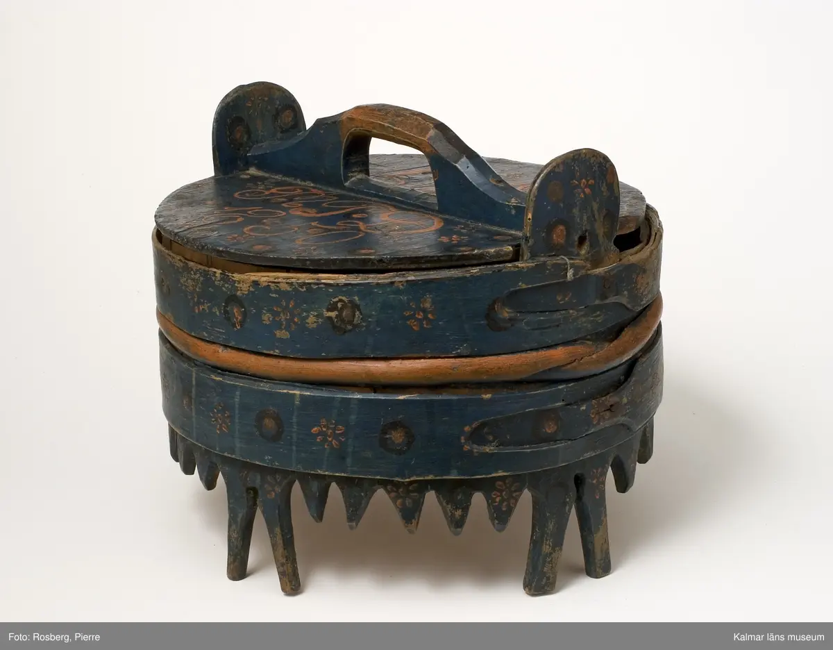 KLM 7627. Grötbytta. Av trä med fyra fötter. Laggad, stavar av bok, lock och botten av furu. Band av spån och vidja. Helt målad. Blå bottenfärg, runt kärlet och på locket röda och svarta blommor och ringar. På locket text: I P S, M P D 1850.