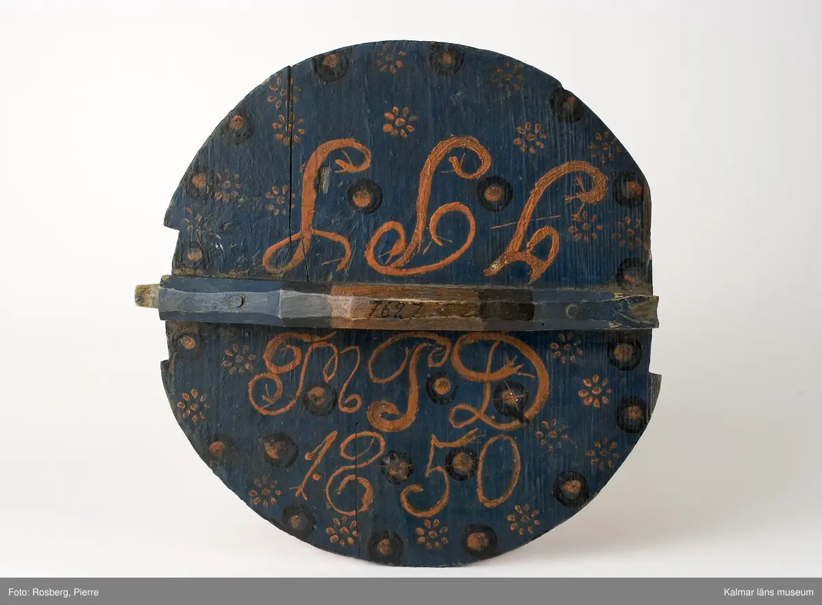 KLM 7627. Grötbytta. Av trä med fyra fötter. Laggad, stavar av bok, lock och botten av furu. Band av spån och vidja. Helt målad. Blå bottenfärg, runt kärlet och på locket röda och svarta blommor och ringar. På locket text: I P S, M P D 1850.