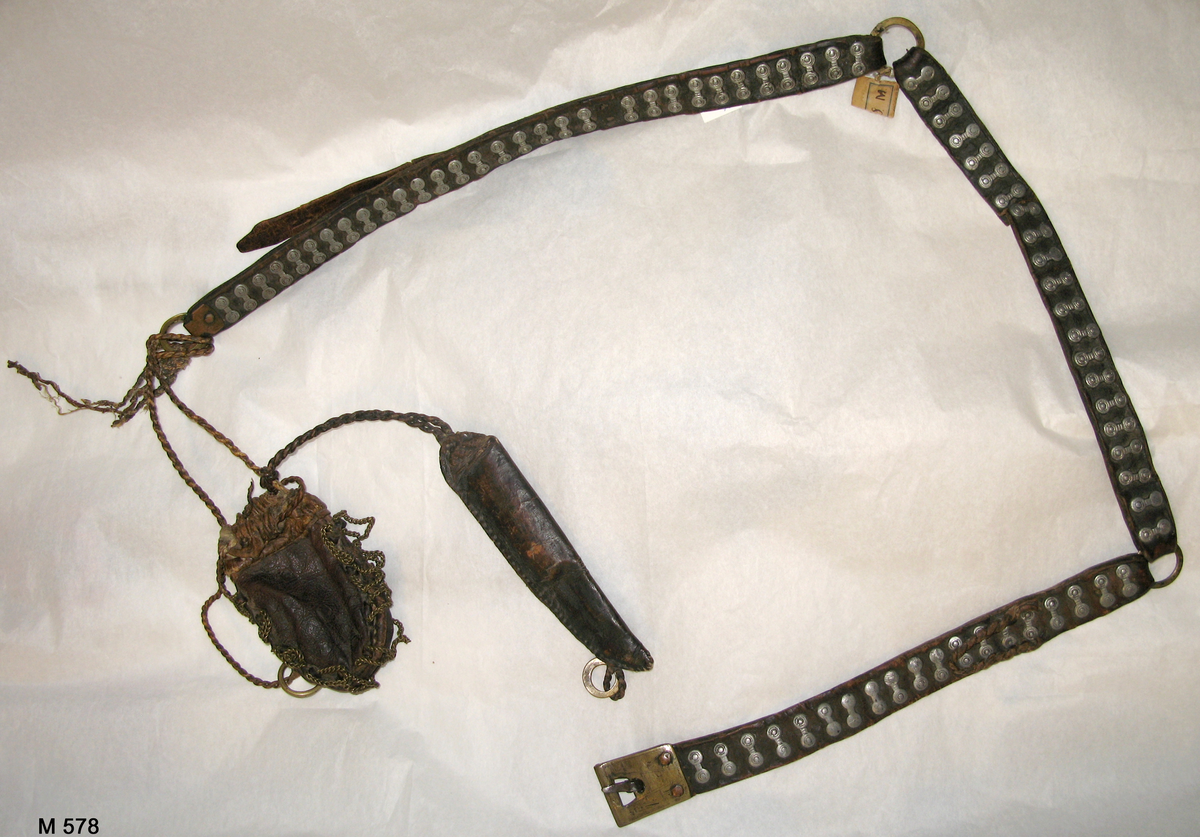 Bälte av tre delar läder hopsatta med två metallringar och med vidhängande pung och knivslida, båda av läder. Bältet är dekorerat med små kläppar av vitmetall (eventuellt tenn) och pungen med mässingskedjor längs sidorna.