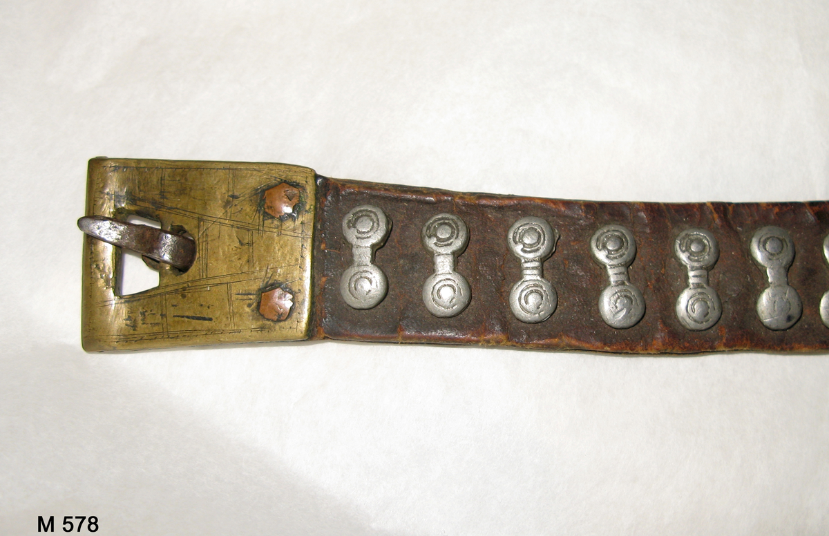 Bälte av tre delar läder hopsatta med två metallringar och med vidhängande pung och knivslida, båda av läder. Bältet är dekorerat med små kläppar av vitmetall (eventuellt tenn) och pungen med mässingskedjor längs sidorna.