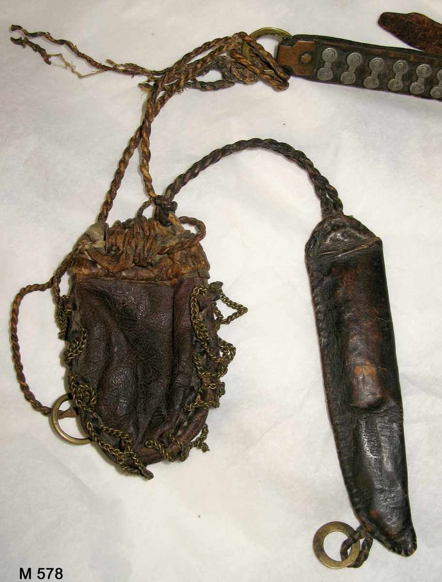 Bälte av tre delar läder hopsatta med två metallringar och med vidhängande pung och knivslida, båda av läder. Bältet är dekorerat med små kläppar av vitmetall (eventuellt tenn) och pungen med mässingskedjor längs sidorna.