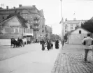 Folkliv på Drottninggatan i höjd med Tyska torget i Norrköping. På höger sida reser sig Grand Hotell, ritat av Werner Northun och öppnat lagom till den publikdragande konst- och industriutställningen 1906. Även byggnaden till vänster var vid tiden tämligen ny, Norrköpings Enskilda Bank uppförd efter Gustaf Wickmans ritningar.