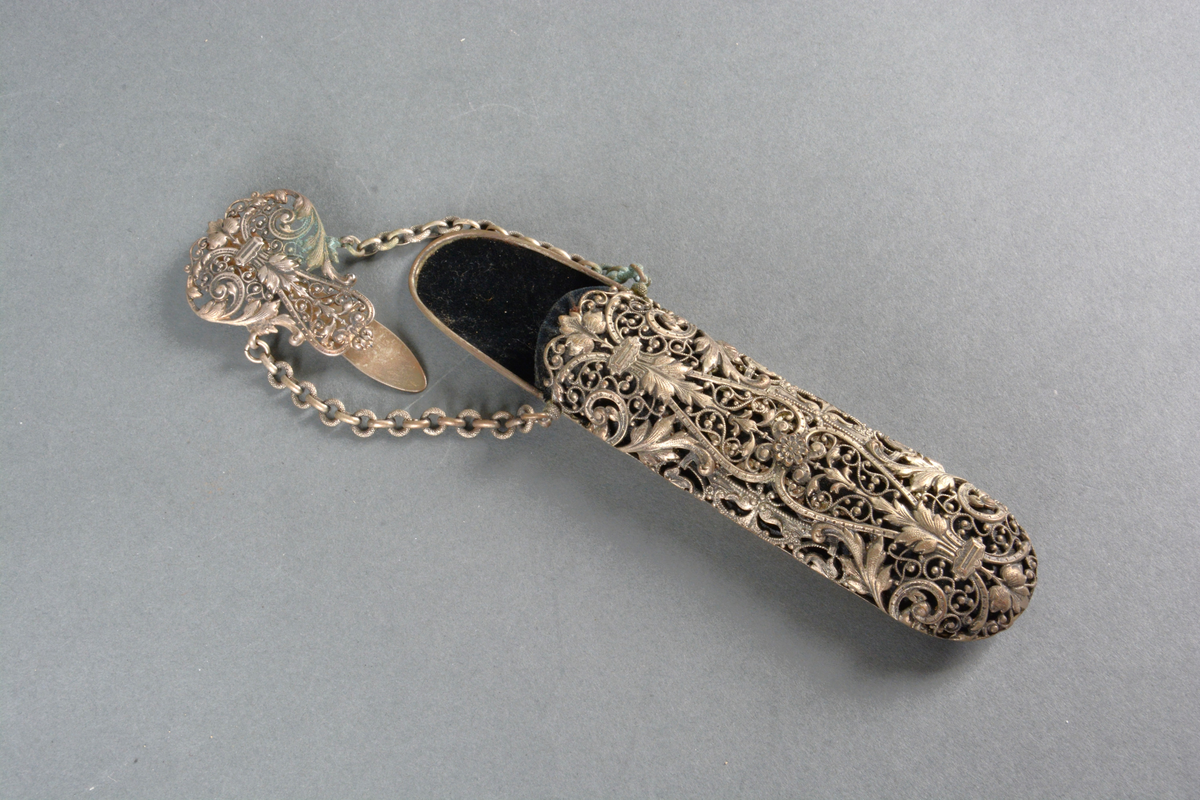 Brille- eller lorgnettfutteral av metall og fløyel. Til å henge i belte. Gjennombrutt rik ornamentikk anbrakt på fremsiden, glatt på baksiden. Foret med sort fløyel. Toppstykket har en hempe til å feste i belte eller skjørtelinning. To kjeder herfra holder selve futteralet.