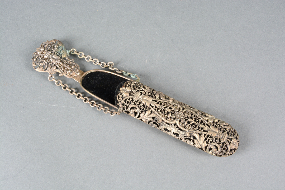 Brille- eller lorgnettfutteral av metall og fløyel. Til å henge i belte. Gjennombrutt rik ornamentikk anbrakt på fremsiden, glatt på baksiden. Foret med sort fløyel. Toppstykket har en hempe til å feste i belte eller skjørtelinning. To kjeder herfra holder selve futteralet.