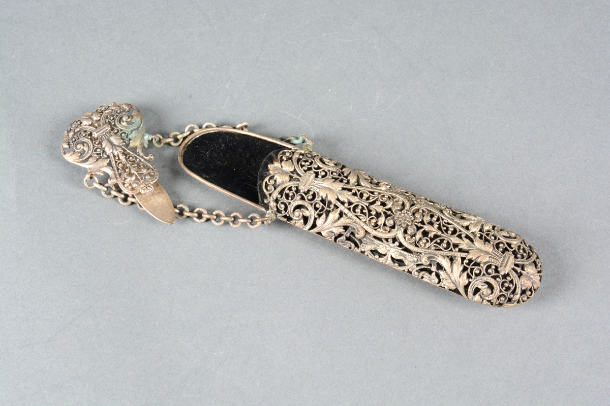 Brille- eller lorgnettfutteral av metall og fløyel. Til å henge i belte. Gjennombrutt rik ornamentikk anbrakt på fremsiden, glatt på baksiden. Foret med sort fløyel. Toppstykket har en hempe til å feste i belte eller skjørtelinning. To kjeder herfra holder selve futteralet.