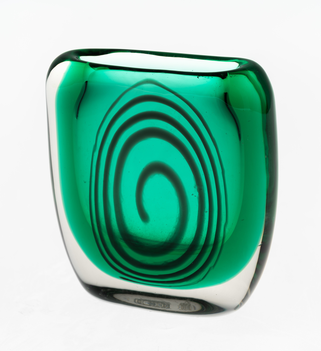 Tilnærmet kvadratisk vase med mykt buede sider i flerfarget glass. Toppen har en oval åpning på midten. Grønnfarget kjerne med blå spiral på en side, omgitt av klart overfang. Planslipt bunn.