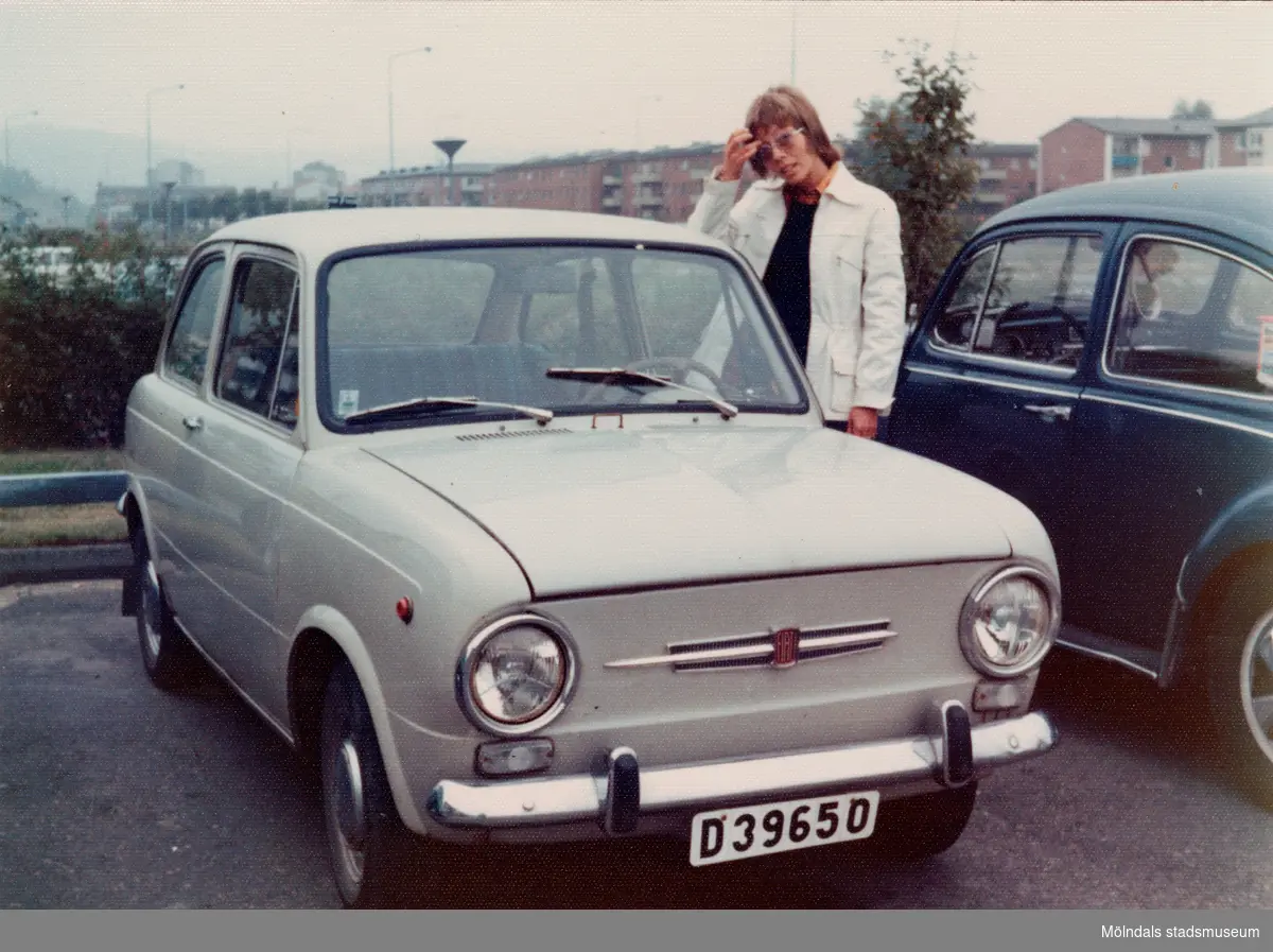 Karin Hansson (född Pettersson) står brevid sin Fiat 650 på parkeringen utanför bostaden på Havrekornsgatan i Bifrost, cirka 1972-1974.