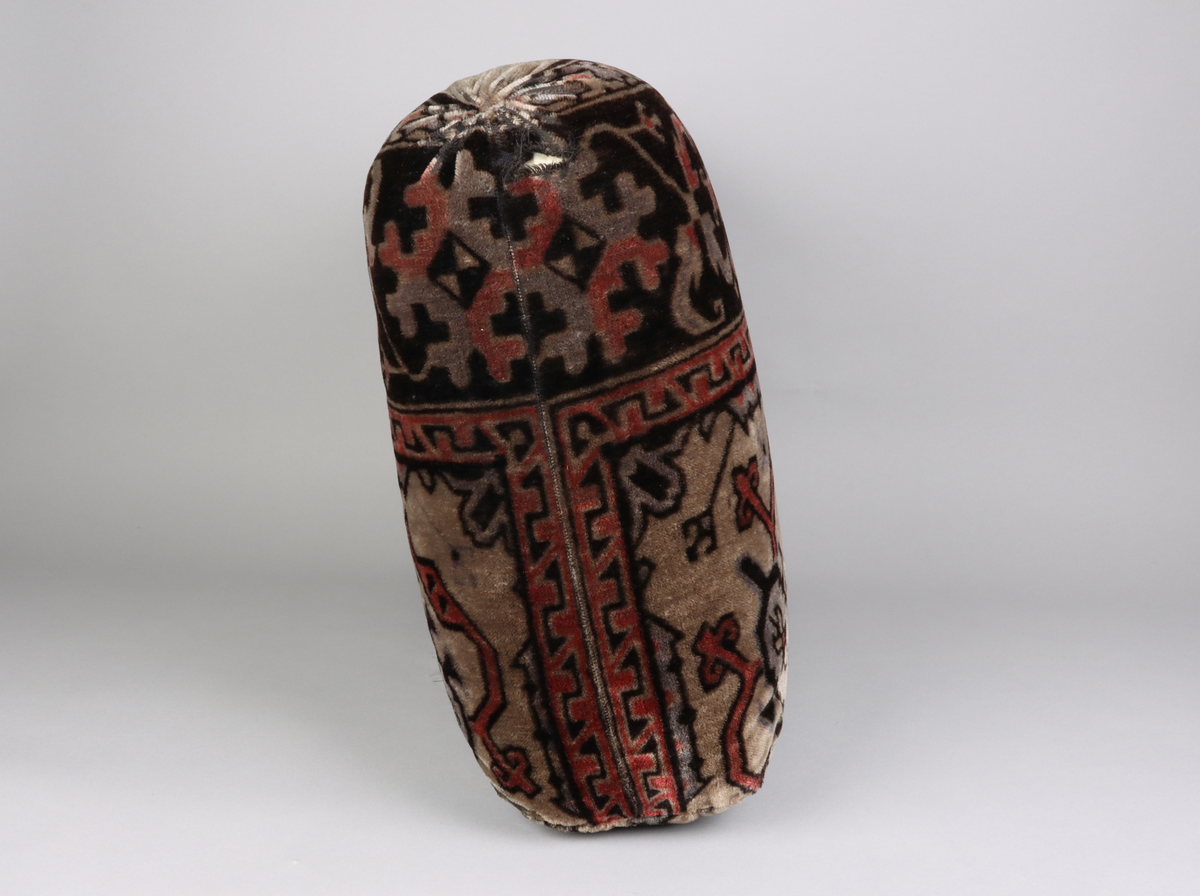 En kudde av mönstrad schagg. Orientaliserande mönster i färgerna grå, svart och vinröd. Del i en grupp med överkast och kuddar till en sk. Ottoman, en typ av bäddsoffa.