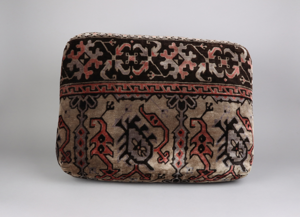 En kudde av mönstrad schagg. Orientaliserande mönster i färgerna grå, svart och vinröd. Del i en grupp med överkast och kuddar till en sk. Ottoman, en typ av bäddsoffa.