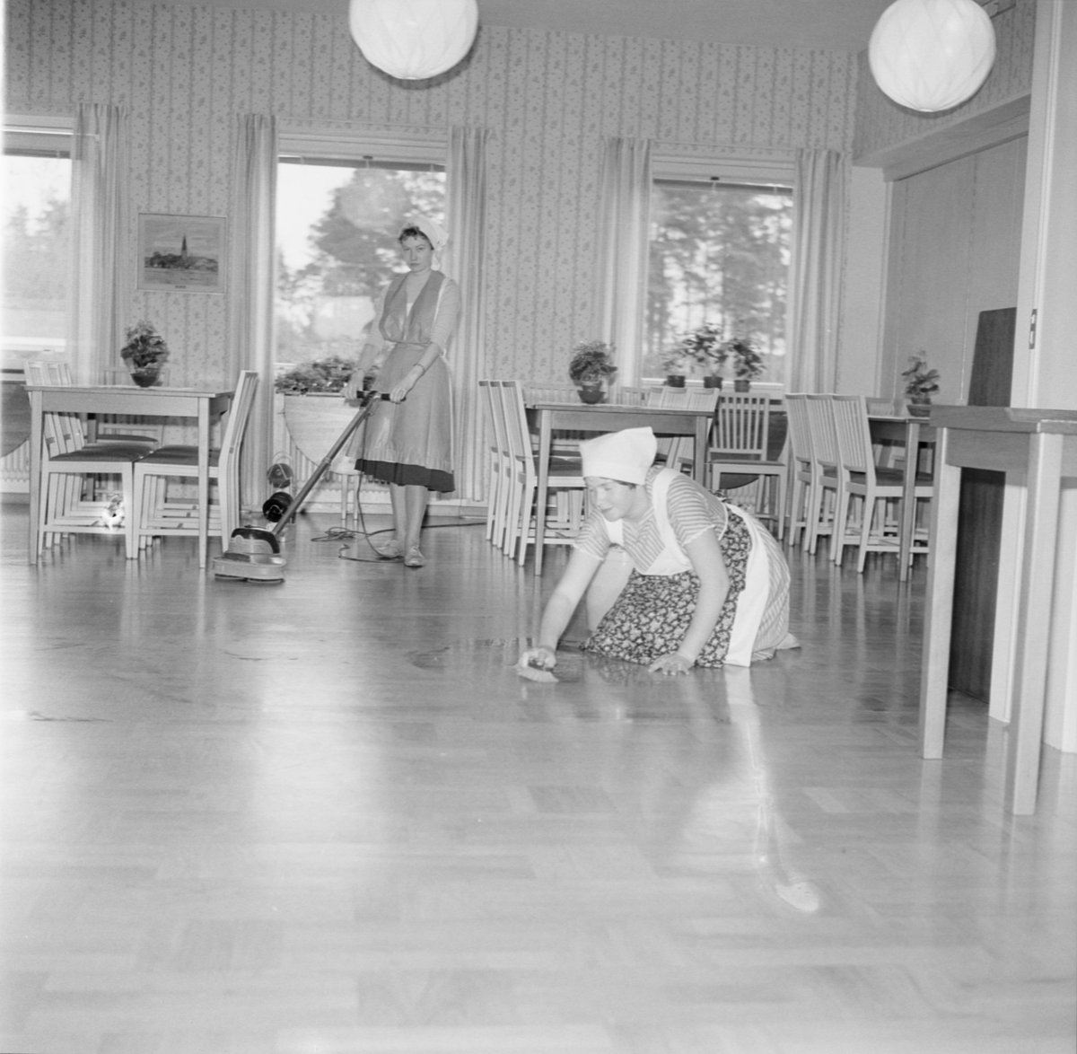 "Fröken Maj-Britt Thorslund skrubbar rent medan fröken Liz Jansson bonar en del av golvet som är klart", sannolikt på Källgårdens Lanthushållsskola