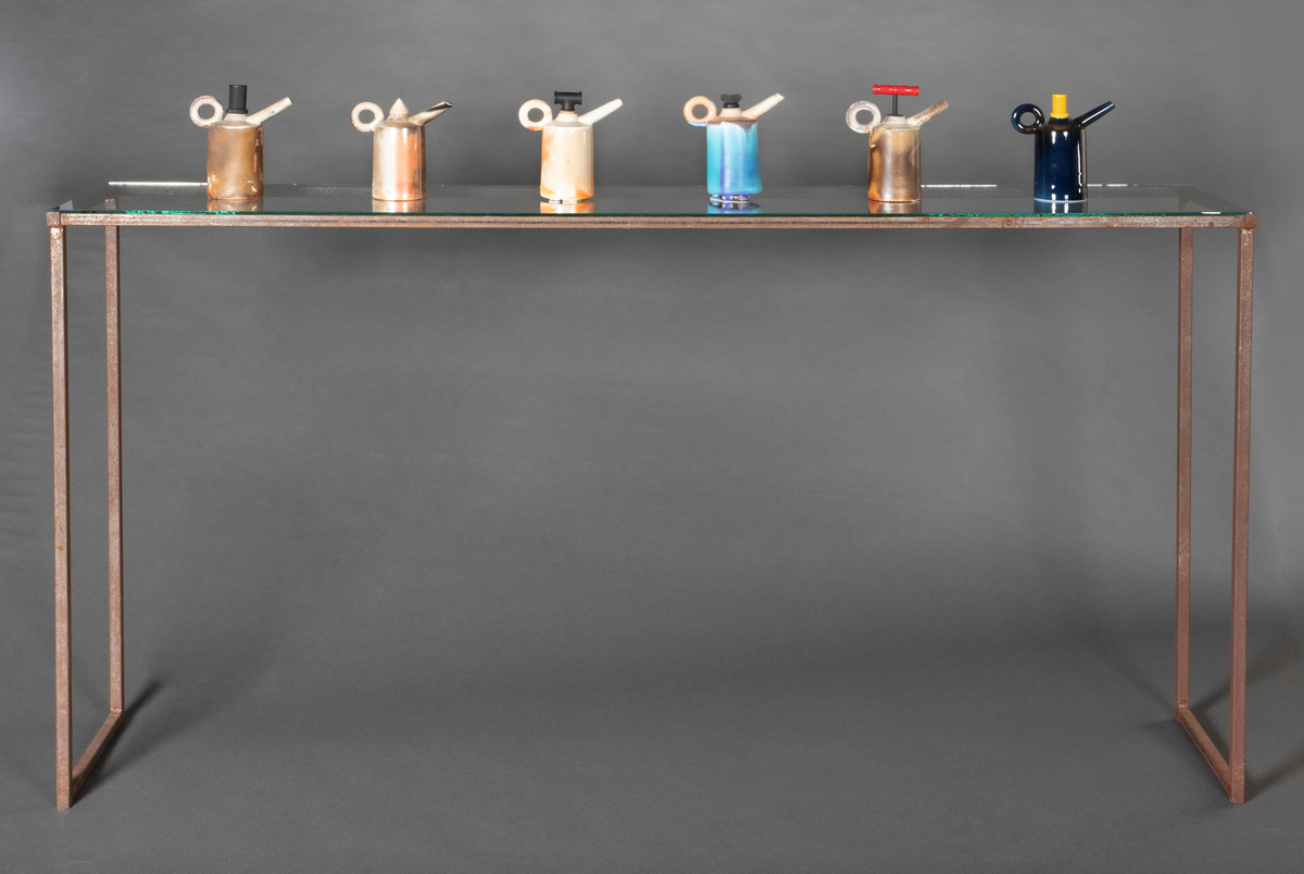 Installasjon med seks polykrome kanner i håndbygd porselen på et rustent bord med glassplate. Kannene er glasert og vedfyrt. Korkene på fem av kannene er lagd av funne komponenter. En har en korkopptrekker, to har industrielle komponenter i lakkert stål og to har gummikomponenter.