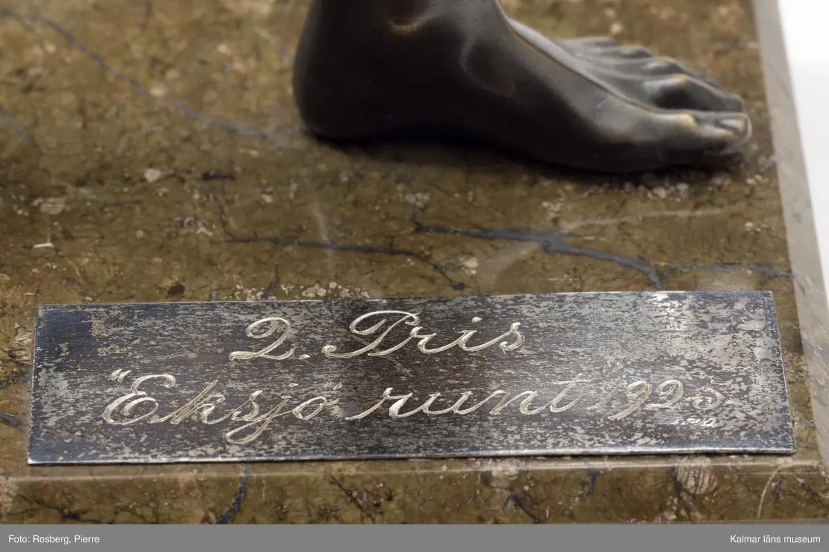 KLM 15376:5. Statyett, av brons och sten. Mansfigur med lagerkvist i uppsträckt hand. Textplatta: 2:a pris Eksjö runt 1925.