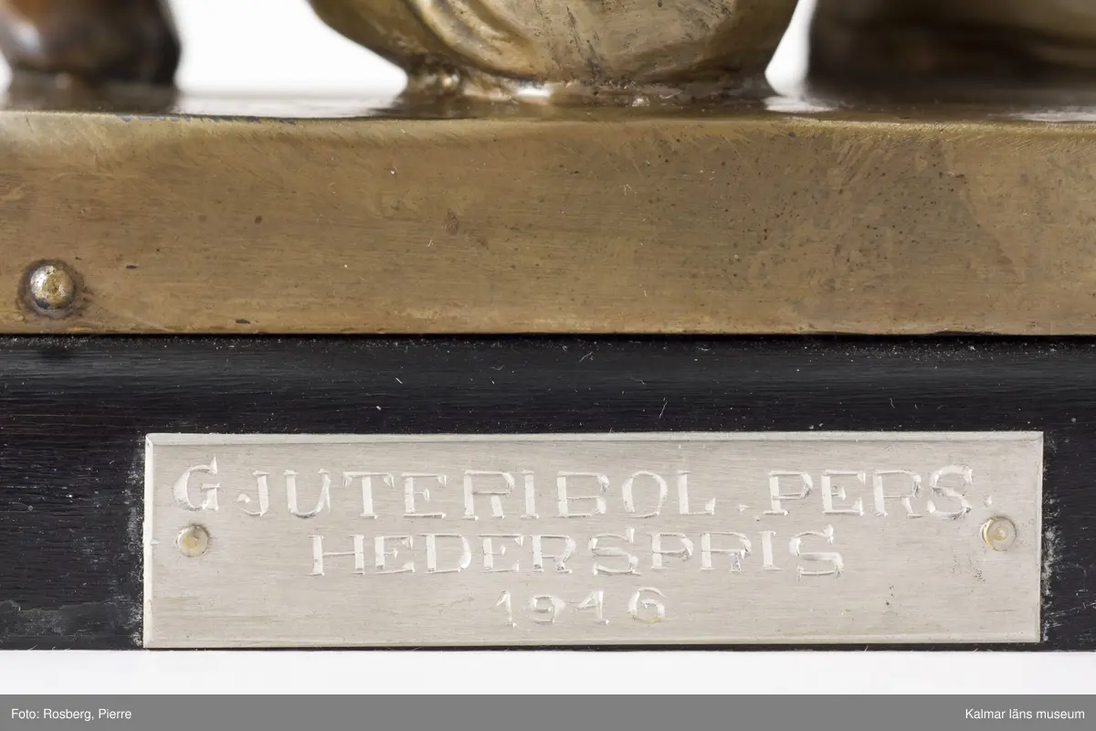 KLM 24282 Statyett, prisstatyett. Statyett av metall, eventuellt brons, i form av en knästående skytt. Plakett i metall med text: Gjuteribol. Pers. hederspris 1946. Påskrift undersida med grön penna: Erövrat av Uno Danielsson, Nils Sallander och Evald Sallander 1946.